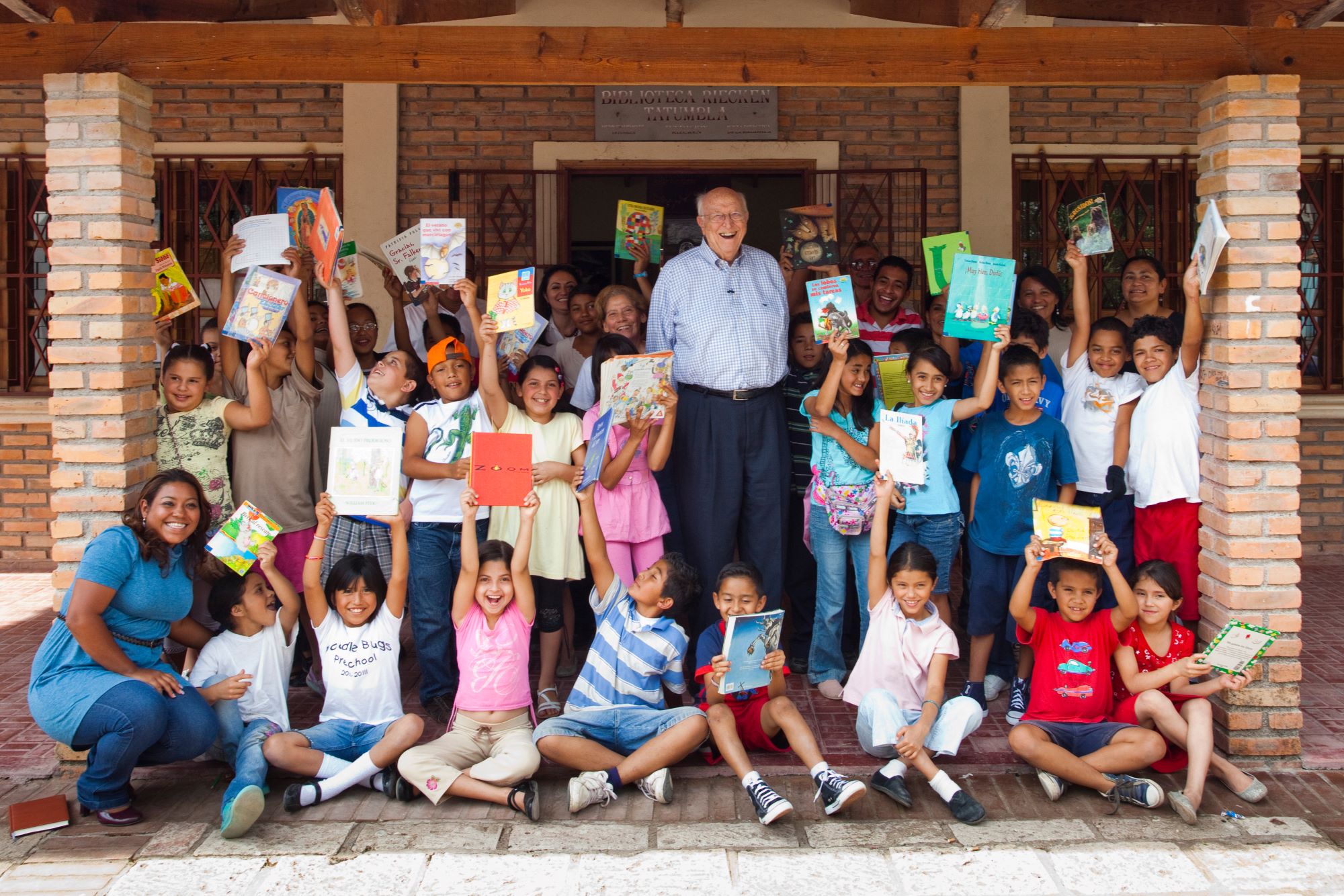 Bill Gates Sr. stands and smiles with a group of young children holding up books smiling in front of the community library in Tatumbla, Honduras.