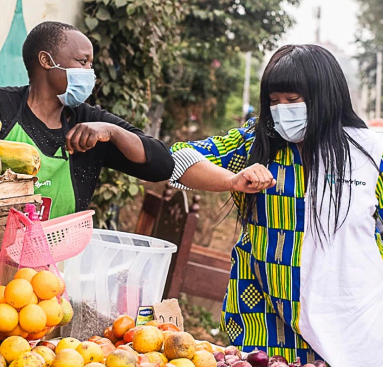 Two people bumping elbows, while wearing masks at a fruit stand.